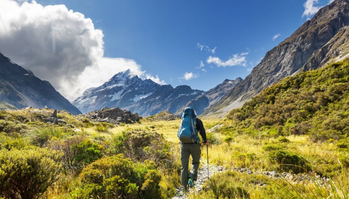 Una persona con mochila camina por un sendero entre montañas, rodeada de vegetación y luz natural.
